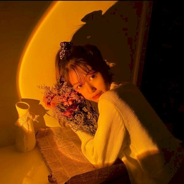 Kiểu ngồi tạo dáng 2 cùi chỏ tựa lên bàn, góc chụp nghiêng từ phía sau lưng, 2 tay ôm bó hoa áp lên má, mắt liếc nhìn ống kính cùng với hiệu ứng đèn vàng