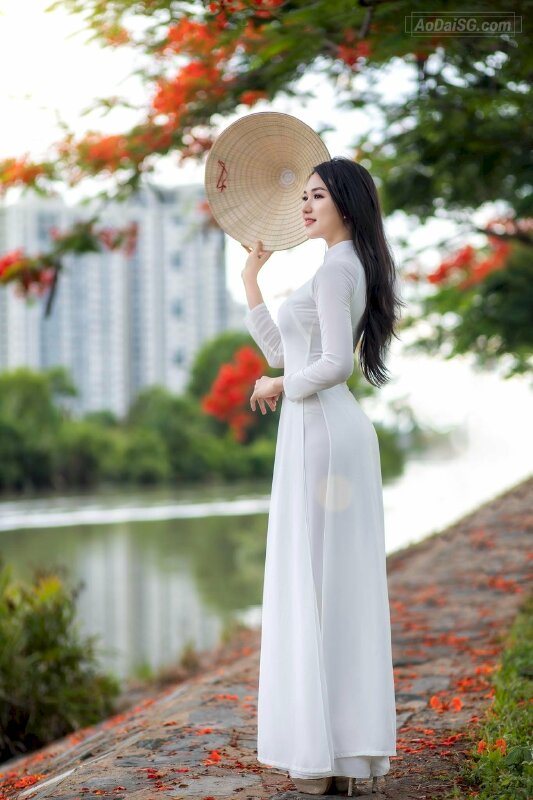 Tạo dáng đứng nghiêng, một tay đưa lên ngang eo, một tay cầm chiếc nón la che nghiêng mặt