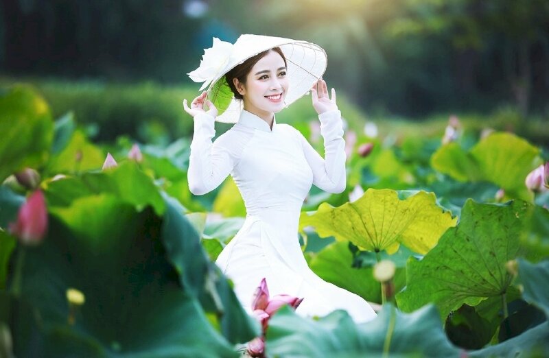 Tạo dáng ngồi dịu dàng, hai tay vịnh nhẹ chiếc nón lá đội trên đầu