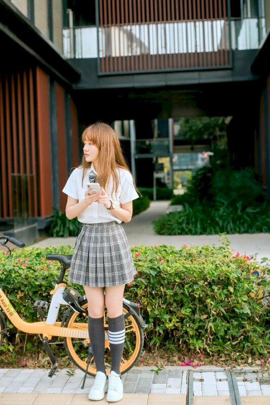  tạo dáng bên cạnh chiếc xe đạp, hai tay cầm điện thoại, mặt hướng nghiêng sang một phía như đang chờ đợi ai đó