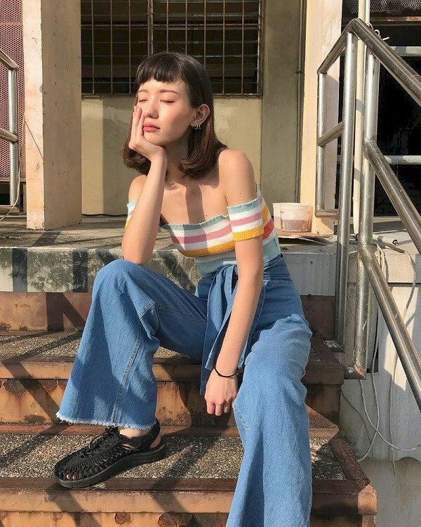 Tạo dáng ngồi trên cầu thang một chân co để trên bậc thang, một chân để thẳng tự nhiên, tay chống cằm