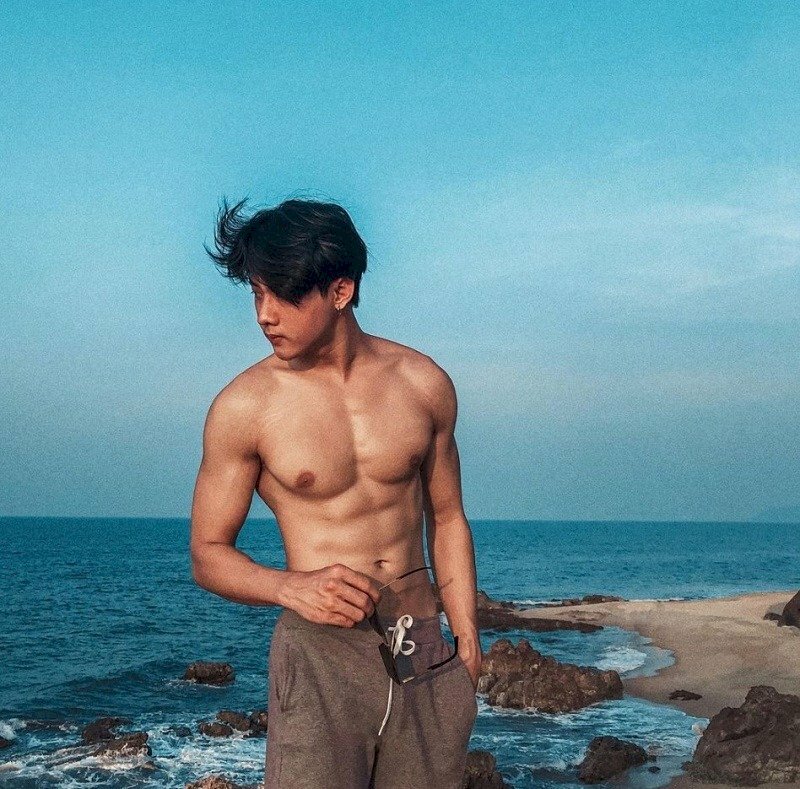 Tạo dáng ở biển với một tay đút túi quần, một tay cầm kính mát, mặt quay nghiêng, tóc bay trong gió