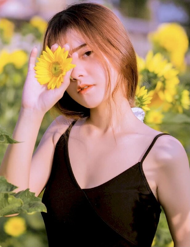 Tạo dáng khi chụp trên cánh đồng hoa cúc vàng với một tay cầm nhanh hoa cúc che một bên mắt phải, đầu hơi nghiêng, mắt nhìn ống kinh