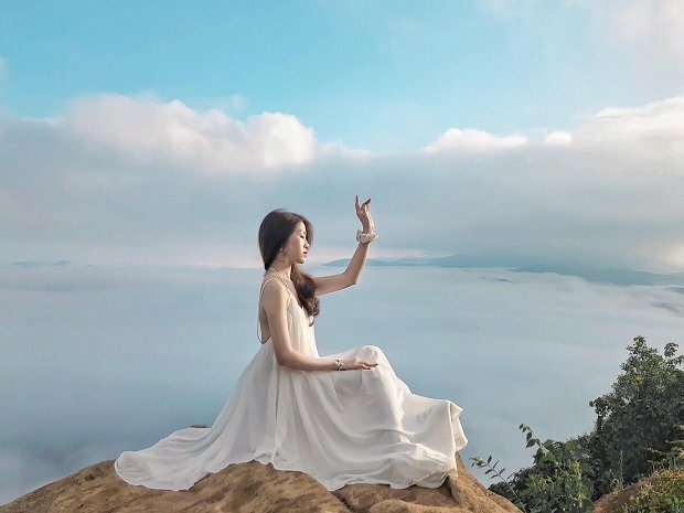 Tạo dáng tư thế ngồi trên một mỏm đá với chiếc đầm phủ kín 02 chân, một tay để tự nhiên trên đùi, một tay giơ lên che nhẹ nhàng