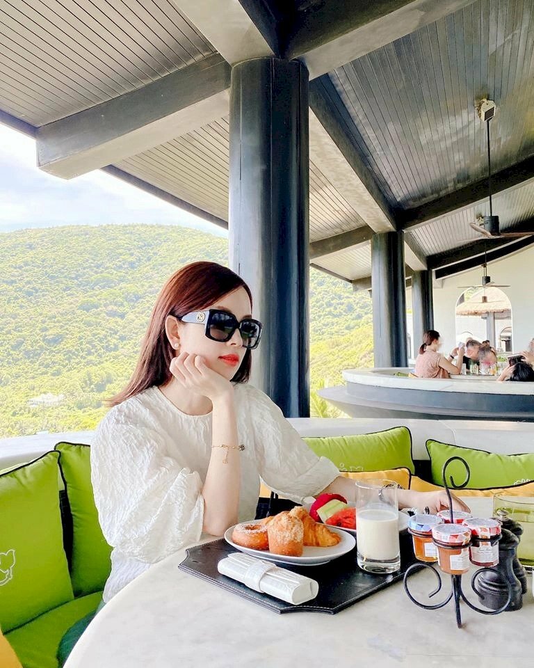 Tạo dáng ngồi bên bàn ăn, một tay chống cằm, một tay để tự nhiên trên bàn, đầu hơi nghiêng với mắt nhìn về phía xa