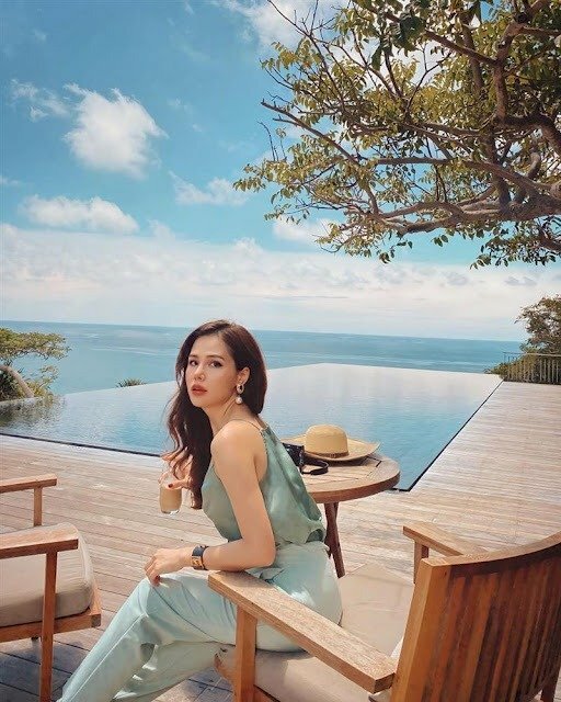 Tạo dáng tư thế ngồi trên ghế hơi khom người về phía trước, hai chân duỗi tự nhiên, một tay đặt lên thành ghế, tóc xoã nhẹ qua một bên, quay lưng và mặt hướng nhìn về ống kính, bờ môi hơi hở một cách quyến rũ