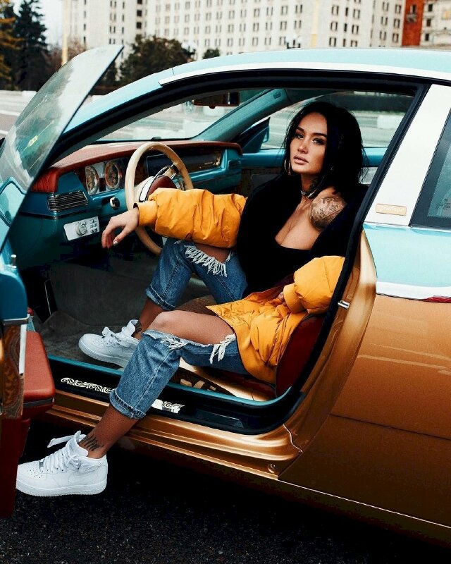 Tạo dáng ngồi trên xe với cửa mở, một chân co gối để tay gác trên xe, một chân duỗi xuống đất, mặt hơi hướng cằm lên nhìn về ống kính