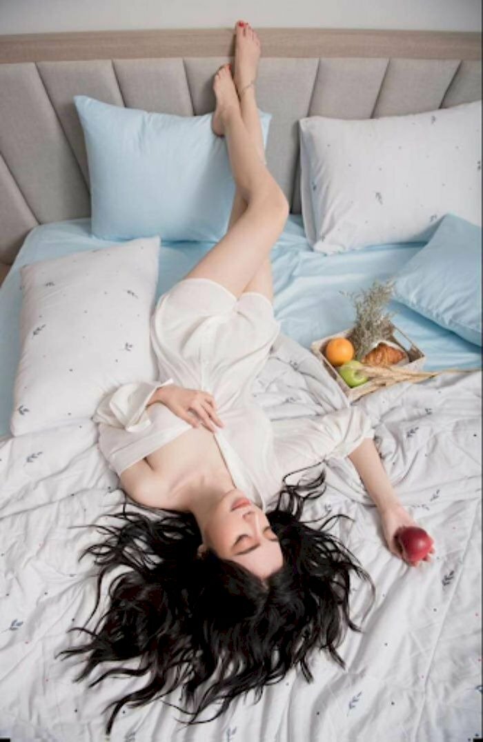 Tạo dáng nằm ngược lại trên giường, hai chân gác lên phía đầu giường tạo thế một chân co, một chân duỗi, một tay vịn áo che phần ngực, một tay để ngửa trên giường cầm một quả táo, đầu nghiêng nhẹ với đôi mắt nhắm