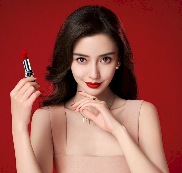 Góc chụp hướng nhìn thẳng ống kính, đầu hơi nhẹ cúi, mắt nhìn hướng ống kính, một tay nhẹ đưa chống cằm, một tay cầm sản phẩm quảng cáo