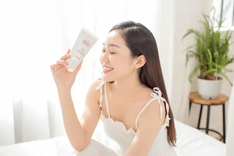 Một chiếc đầm trắng 02 dây nhẹ nhàng, tạo dáng ngồi nghiêng, tóc dài xoã ngang lưng, miệng cười, mắt nhắm khẽ, một chân co gối để cánh tay cầm sản phẩm chống nhẹ nhàng và cầm sản phẩm đưa nhẹ hướng vào gương mặt