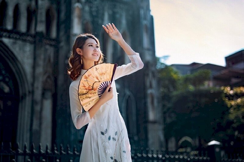 Tạo dáng đứng thật tự nhiên với mặt hướng nhìn sang một phía, đưa nhẹ tay lên che nắng, tay kia có thể cầm phụ kiện là chiếc quạt đưa lên ngang ngực