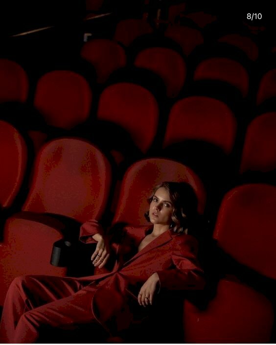 Tạo dáng ngồi cá tính của cô gái trong bộ quần áo màu đỏ giữa hàng ghế trống trong rạp chiếu phim với tư thế ngồi tựa người dài trên ghế, 2 tay gác lên, mắt nhìn ống kính biểu cảm mệt mỏi