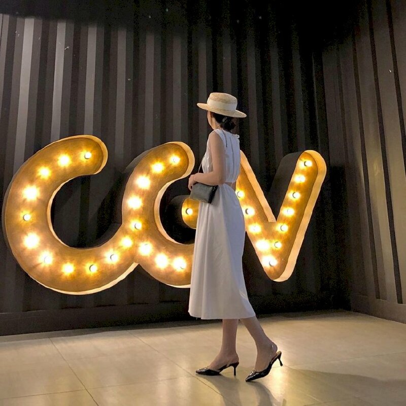 Tạo dạng chụp ảnh tại rạp CGV trong chiếc váy trắng dịu dàng, đầu đội nón vành cùng tư thế đứng quay ngưới với góc chụp từ phía sau hơi nghiêng, vai mang túi, tay đặt lên chiếc ví đen xinh xắn, một chân bước lui về sau thể hiện sự thướt tha đầm thấm