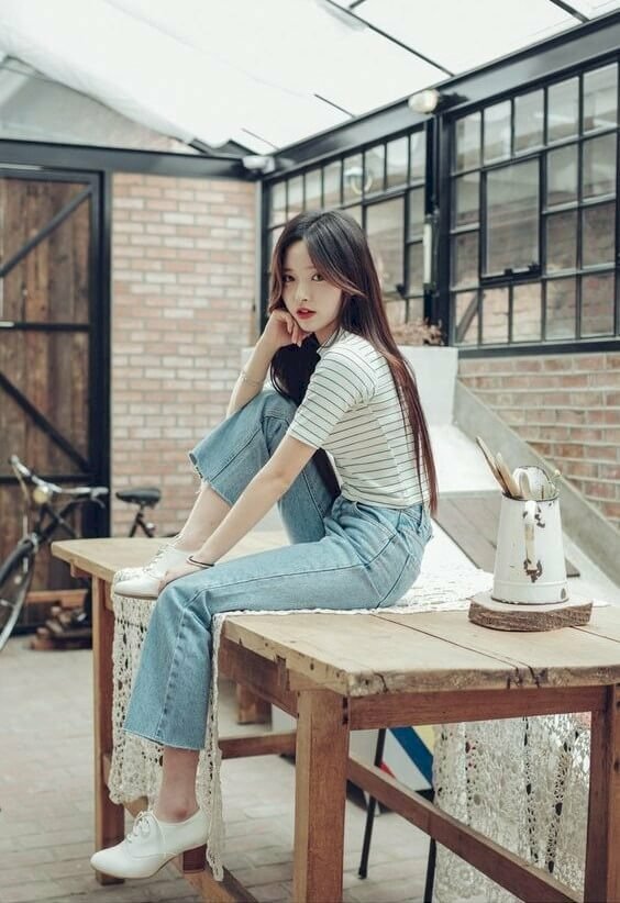 Mẫu tạo dáng ngồi trên một cái bàn gỗ, một chân co, tay chống cằm, chụp ảnh cá tính thu hút
