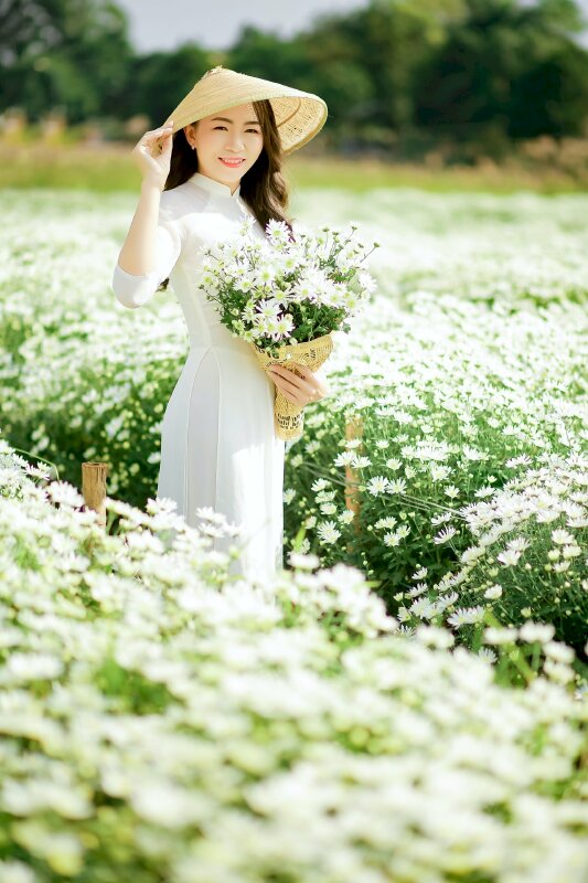 Cách tạo dáng với hoa trong bộ áo dài trắng, một tay cầm bó hoa cúc trắng, một tay cầm lấy vành nón lá che nắng, chụp góc nghiêng, mặt hướng về ống kính