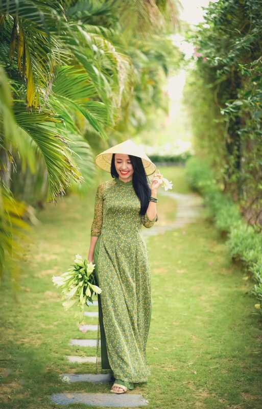 Kiểu ảnh trong bộ áo dài xanh đang bước đi thong dong miệng mỉm cười, 1 tay cầm bó hoa ly, 1 tay chạm vành nón lá kèm một nhánh hoa nhỏ tuyệt đẹp
