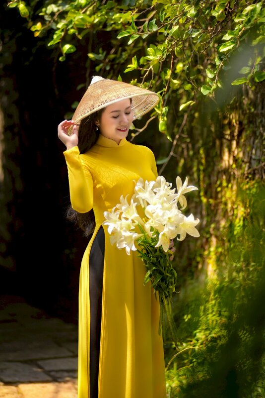 Chụp ánh trong bộ áo dài vàng, dáng người đứng nghiêng, 1 tay cầm bó hoa ly, 1 tay chạm vành nón lá, đầu hơi nghiêng, mắt nhìn xuống bó hoa ly