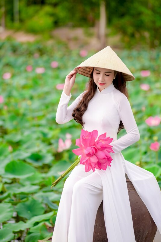 Tạo dáng trong bộ áo dài trắng tinh khiết, đang tựa vào thành chiếc lu nước, 1 tay cầm bó hoa sen hồng thắm, 1 tay nắm vành nón lá, đầu hơi cúi, mắt nhìn xuống bó hoa