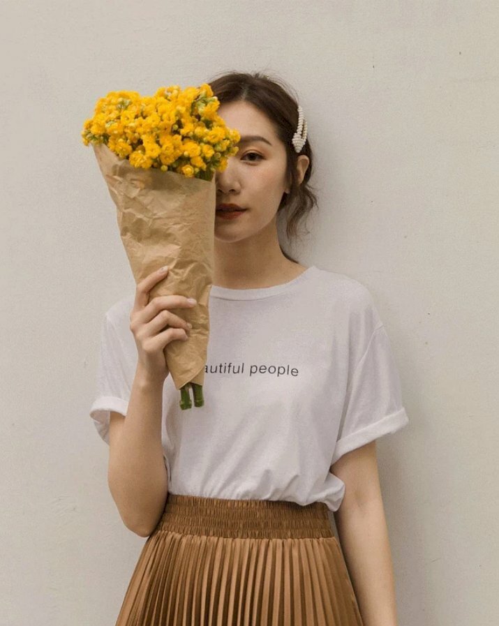 Tạo dáng đẹp thanh thoát trong chiếc váy nâu và áo thun đơn giản cùng tay cầm bó hoa cúc vàng baby che nửa mặt bên phải, tay còn lại để xuôi tự nhiên