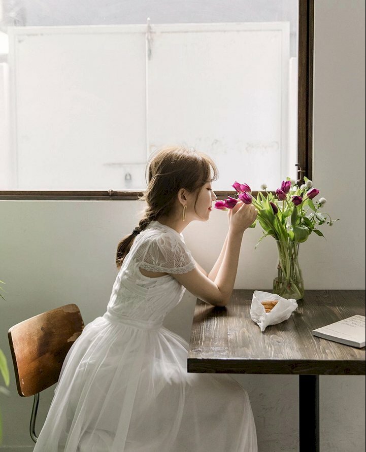 Kiểu chụp ảnh với hoa để phòng trong bộ đầm trắng, dáng ngồi hơi chòm về phía bình hoa, tay nắm lấy nhanh hoa, mặt diễn nét chill phết với hương hoa tím, góc chụp nghiêng trông dễ thương