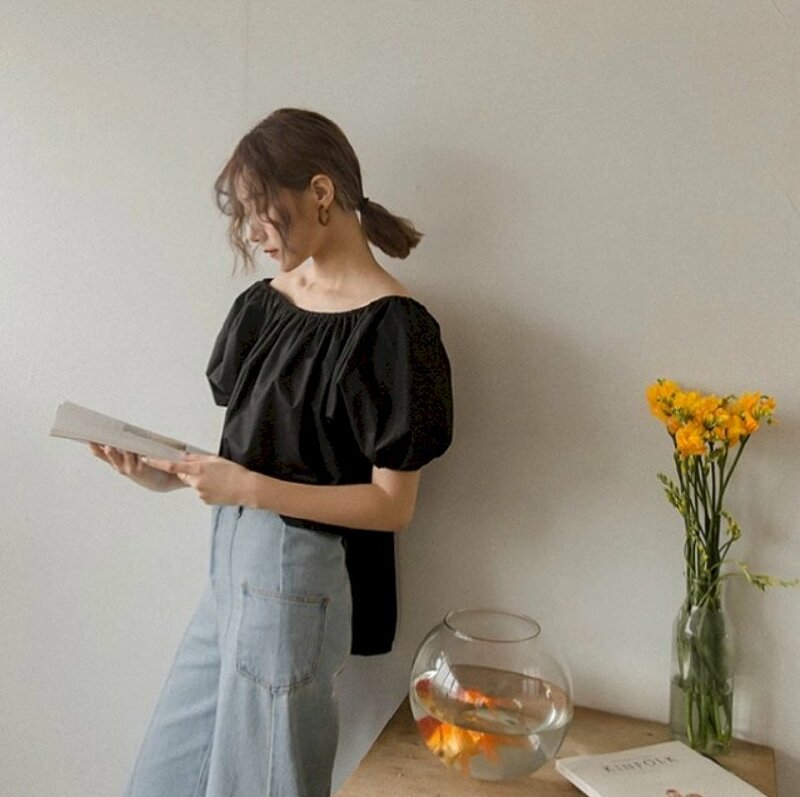 Tạo dáng chụp ảnh đẹp trong phòng với kiểu đứng tự nhiên tựa lưng vào tường, 1 chân hơi co, 2 tay cầm sách, mắt hơi cúi nhìn sách, cùng bình hoa vàng chưng tuyệt đẹp bên chậu cá sễ thương