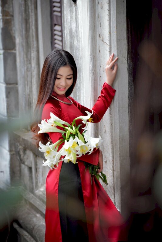 Tạo dáng chụp ảnh với áo dài góc nghiêng, một tay vịnh nhẹ bờ tường, một tay ôm bó hoa đẹp, đầu nghiêng hơi cúi nhìn hoa với miệng cười đẹp, tóc dài xoã
