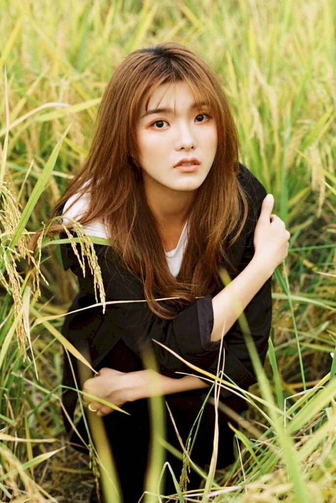 Tạo dáng ngồi giữa cánh đồng lúa với tạo dáng ngồi co gối, một tay ôm gối, một tay ôm nhẹ bờ vai, mặt hướng nhìn ống kính, cằm đưa nhẹ hướng lên trên