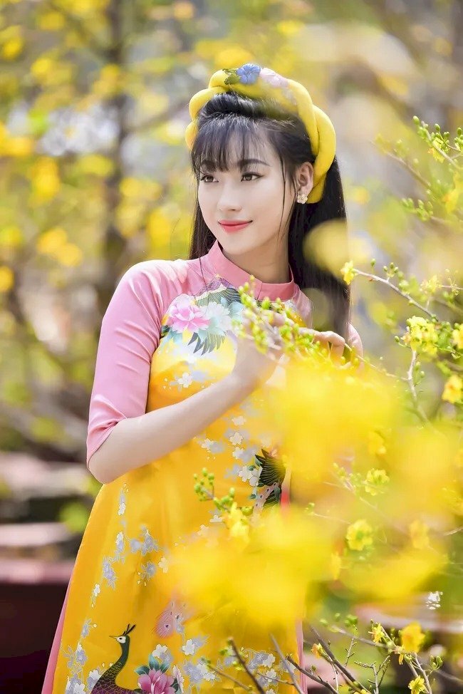 Tạo dáng đứng thật tự nhiên bên hoa mai, hai tay vịnh nhẹ lên cành hoa, dáng người hơi nghiên về phía hoa, mặt nhìn không hướng về máy ảnh