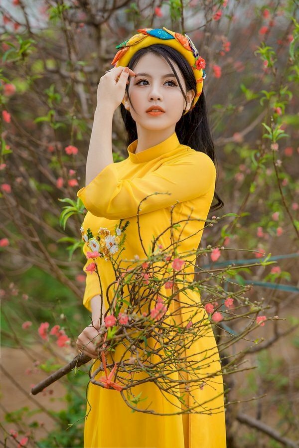 Tạo dáng đứng nghiêng với một tay cầm cành đào, một tay nhẹ đưa lên vuốt tóc, mắt nhìn về phía xa