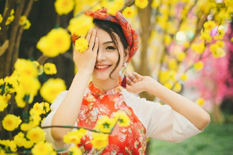 Tạo dáng một tay che nửa mặt với một bông hoa mai kẹp giữa các ngón tay, tay kia thật tự nhiên hất lên trên và ra phía sau