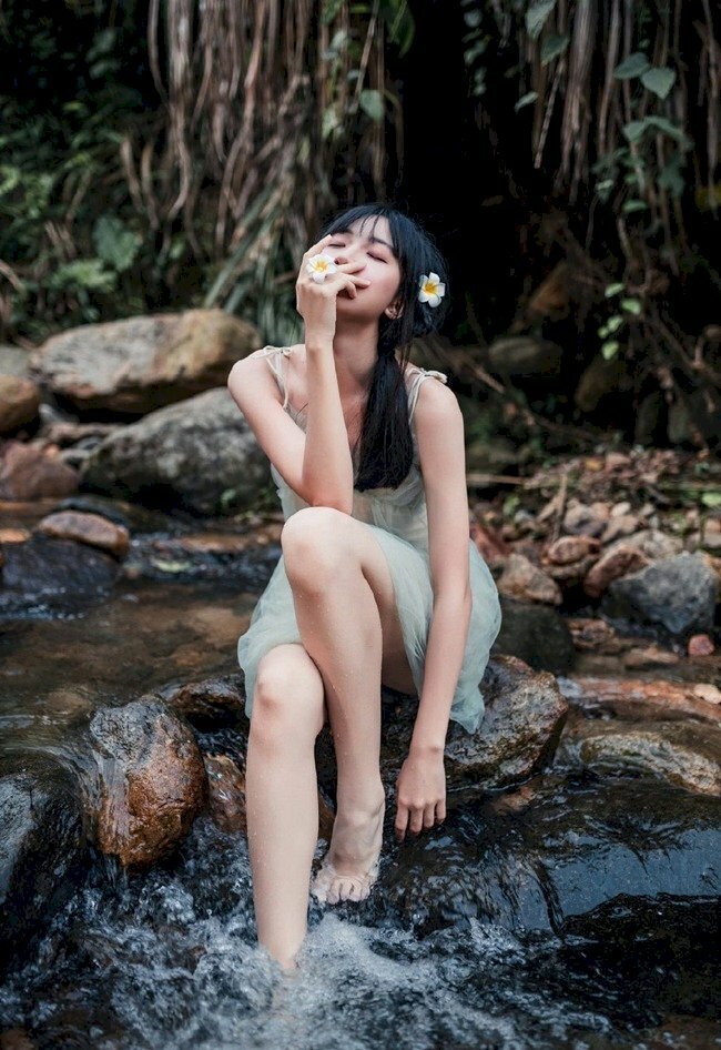 Tạo dáng chụp ảnh ngồi trên mỏm đá, một chân co gác lên đá, một chân duỗi tự nhiên xuống suối, một tay buông tự nhiên, một tay nhẹ đưa lên che mặt, mặt hướng nhìn lên trên với mắt nhắm nhẹ nhàng