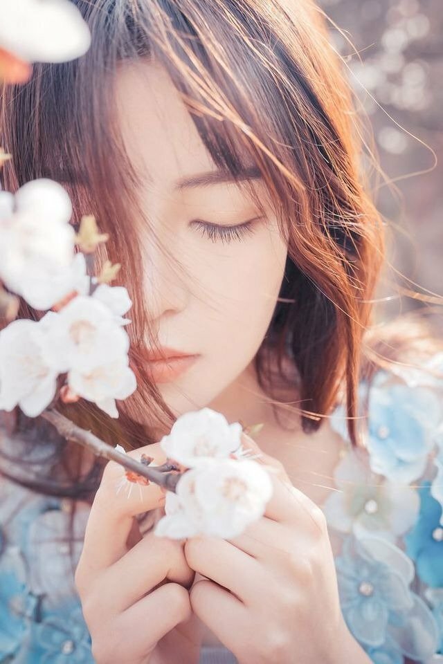 Góc chụp ảnh chính diện, gương mặt thanh tú của bạn gái hướng về máy ảnh với đôi mắt nhắm nhẹ hướng xuống, hai tay cầm một cành hoa che nửa gương mặt