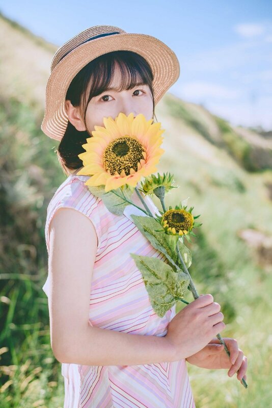Góc chụp nghiêng với tạo dáng đứng tự nhiên, mặt hướng nhìn ống kính, hai tay cầm một cành hoa hướng dương che một phần gương mặt để lộ đôi mắt ngây thơ