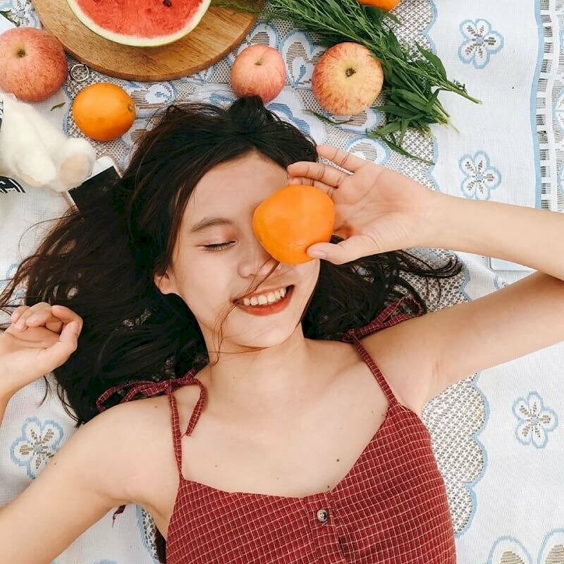 Tạo dáng dùng tay cầm một quả cam, quả táo... che đi một bên mắt, miệng cười thật tươi, mắt còn lại khép nhẹ
