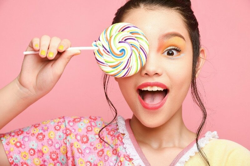 Tạo dáng chụp ảnh che một phần mặt với một chiếc kẹo mút đủ màu, nét mặt tươi vui với khuôn miệng há to, mắt mở to dễ thương