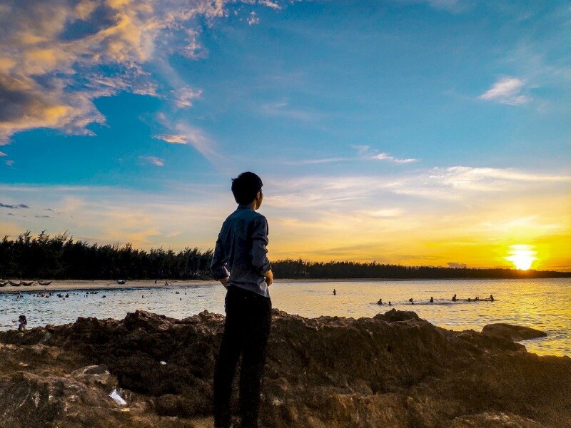 Chàng trai đứng góc nghiêng quay lưng về phía ống kính, hai tay đút túi quần, mặt hướng nhìn ra biển