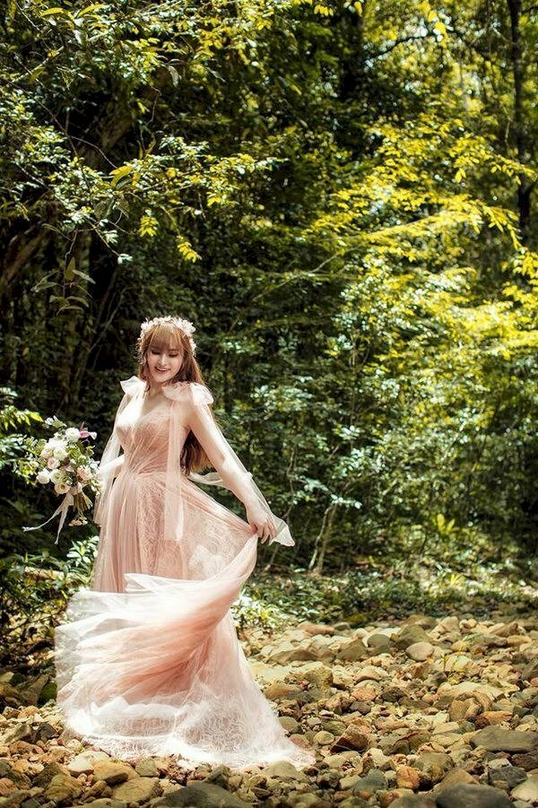 Một chiếc đầm dài thướt tha, một vòng hoa trắng xinh đội đầu, tạo dáng như đang xoay người với một tay nhẹ cầm váy, một tay cầm một bó hoa dại, mặt hơi cúi nhẹ