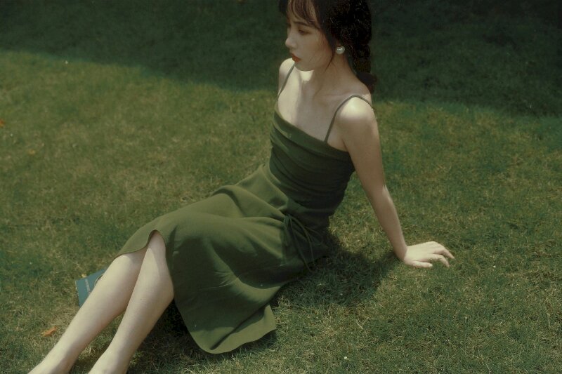 Tạo dáng ngồi trên bãi cỏ với 02 chân duỗi nhẹ, 02 tay chống ra sau, gương mặt biểu cảm thoáng nét buồn