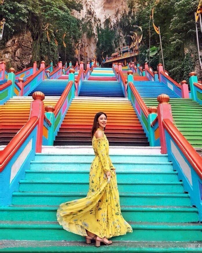 Tạo dáng đứng nghiêng người trên cầu thang, mặt hướng về ống kính với nụ cười tươi, tay cầm nhẹ váy