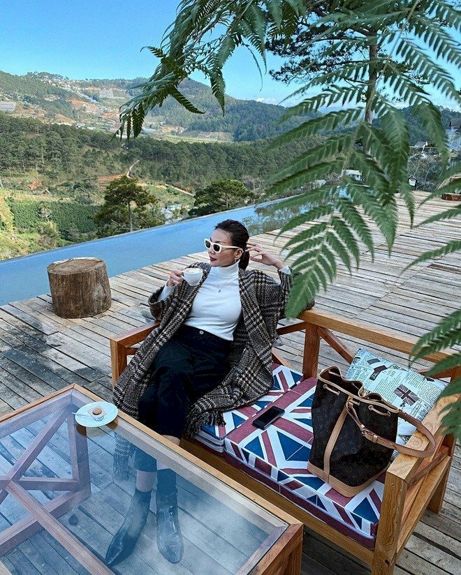 Tạo dáng tư thế ngồi ngả tựa lưng vào ghế, một tay gác lên thành ghế phía sau chống nhẹ vào đầu, một tay gác trỏ lên tay vịn ghế cầm ly cà phê, hai chân bắt chéo hơi duỗi, mặt quay nghiêng