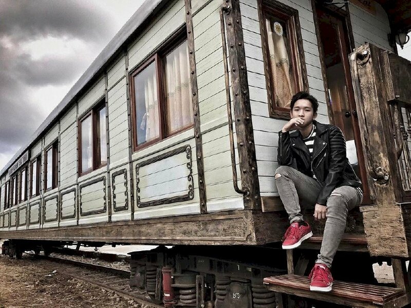 Tư thế tạo dáng ngồi 02 chân co để trên và dưới, người hơi ngả dựa vào thành toa xe, một tay chống trỏ lên đầu gối che ngang miệng, mắt nhìn vào ống kính