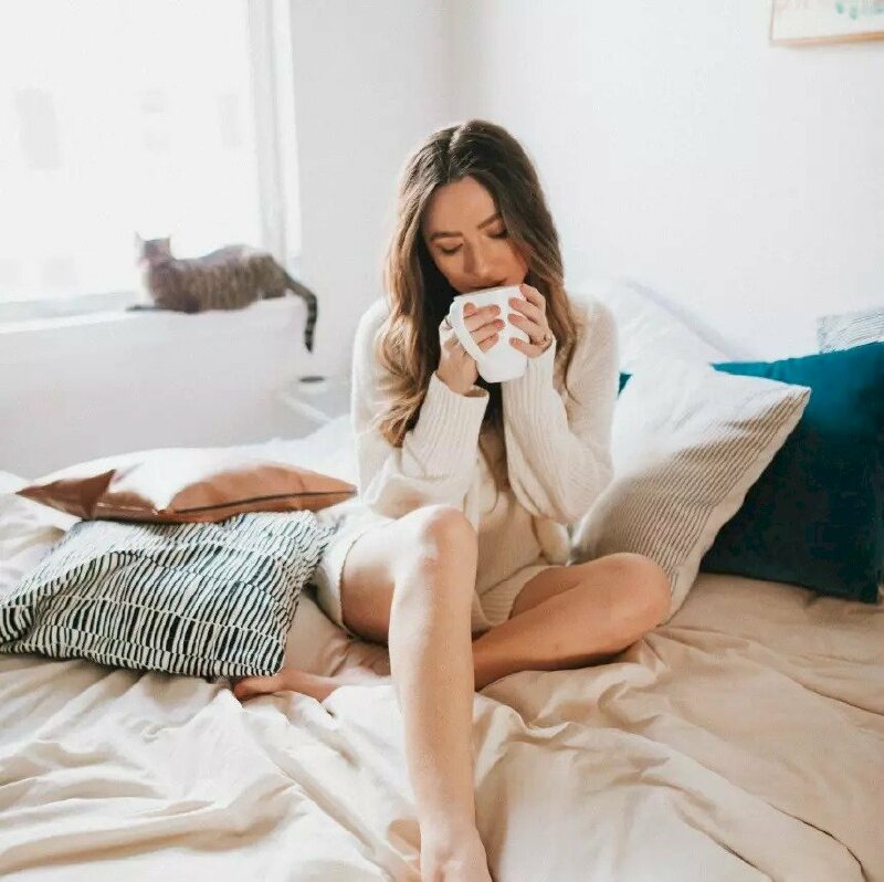 Tạo dáng ngồi trên giường thật thoải mái, một chân xếp ngang, một chân co gối, hai tay nâng tách cà phê, mặt hơi cúi