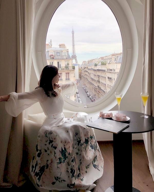 Thả dáng ngồi trên ghế, mặt quay về hướng cửa sổ, hai tay dang rộng sang ngang, góc chụp nghiêng