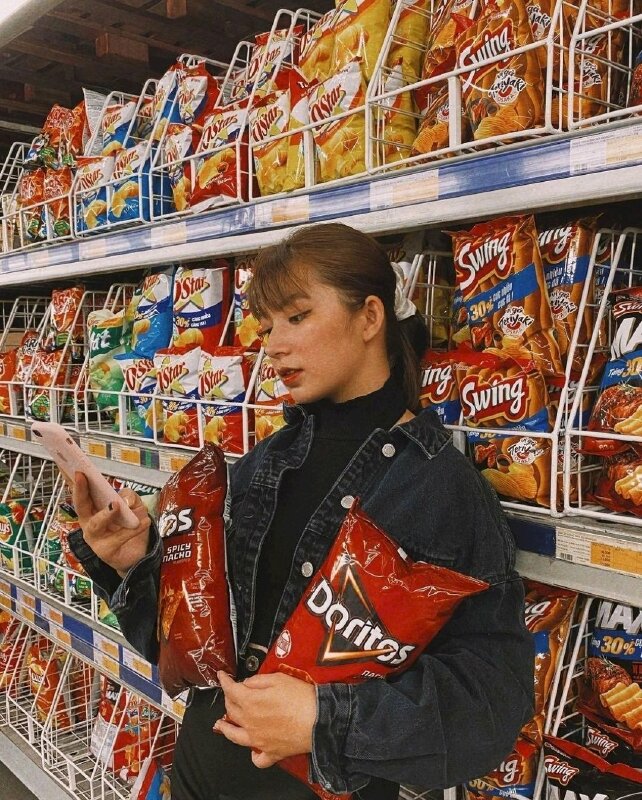 Tạo dáng đứng tựa nhẹ người vào quầy hàng bánh snack, ôm hai gói bánh snack màu sắc rực rỡ, một tay giả vờ như đang bấm điện thoại