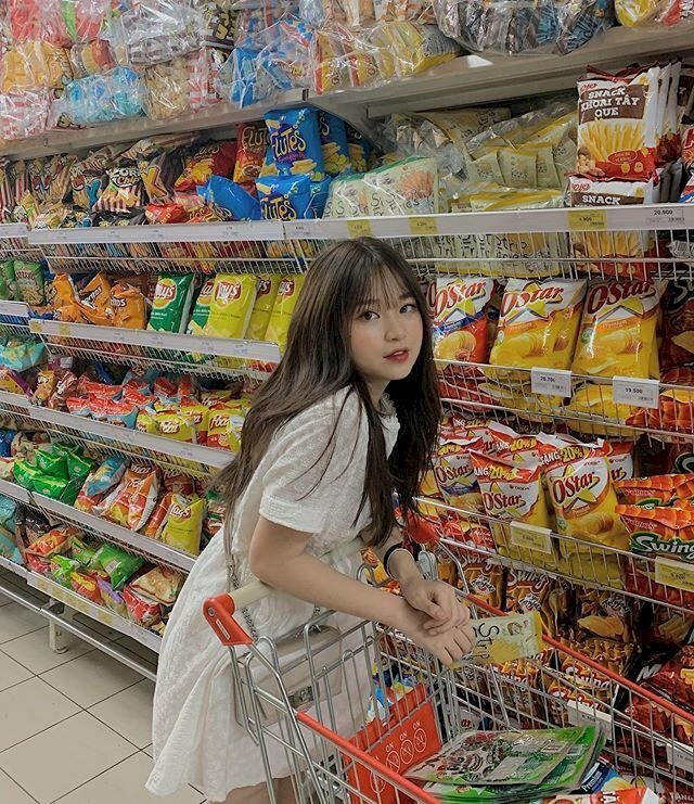 Tạo dáng hạ người nhẹ nhàng tựa vào xe đẩy hàng, hai tay chống hờ lên tay vịnh của xe đẩy, một bên vai khẽ hếch lên trên, mặt quay nhìn vào ống kính với đôi mắt to tròn đáng yêu
