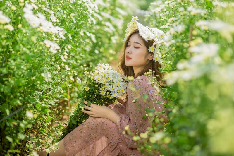 Tạo dáng ngồi nghiêng khẽ co gối, khuôn mặt quay hướng về ống kính, cằm khẽ hướng lên trên, mắt nhắm nhẹ mơ màng, tay ôm bó cúc hoạ mi