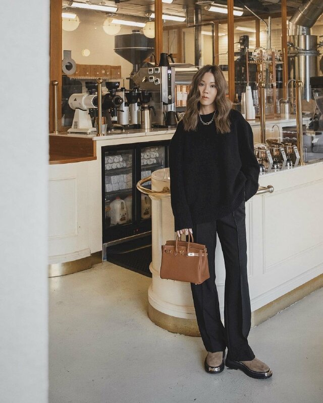 Tạo dáng đứng tựa hông, một chân trụ thẳng, một chân với mũi chân hơi hướng nghiêng, một tay cầm túi buông thõng, tay còn lại đút túi quần