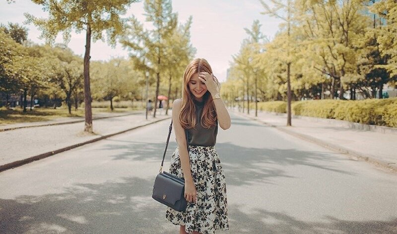 Tạo dáng đứng tự nhiên với đầu cúi nhẹ cười thật duyên, một tay nhẹ đưa vuốt tóc, một tay buông thẳng tự nhiên với một chiếc túi thời trang đeo trên vai