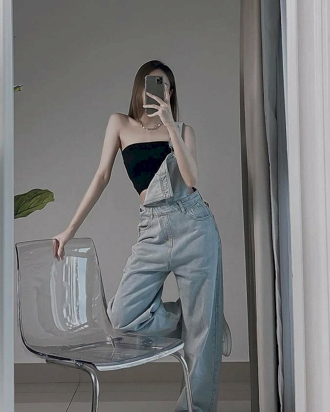 Tạo dáng chụp trước gương với ghế, một chân co gác lên ghế, một chân đứng thẳng tự nhiên, một tay vịn ghế, một tay cầm điện thoại che mặt