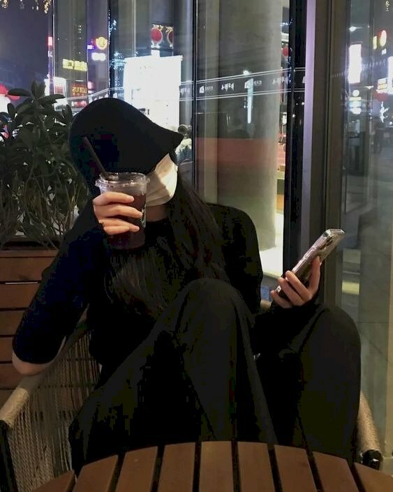 Tạo dáng ngồi co chân trên ghế, thân người dựa vào ghế, một tay gác lên thành ghế cầm ly trà sữa che mặt